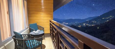 A balcony with a wooden railing, a table with chairs, and a view of a starry night sky and distant town lights.