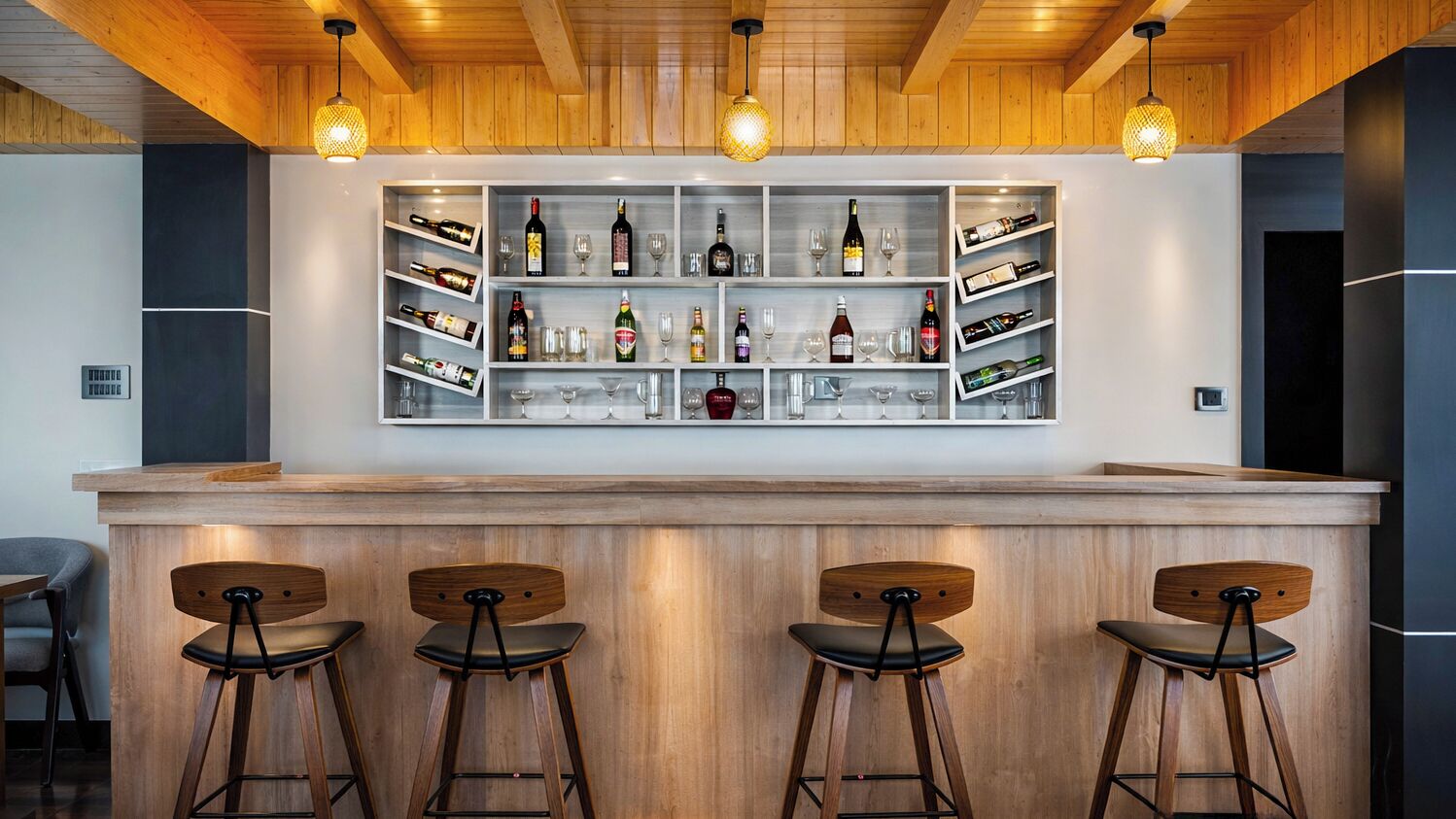 Un área de bar con taburetes de madera, estantes con botellas y copas, e iluminación colgante.
