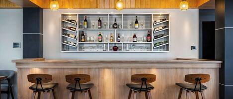 A bar area with wooden bar stools, shelves with bottles and glasses, and pendant lighting.