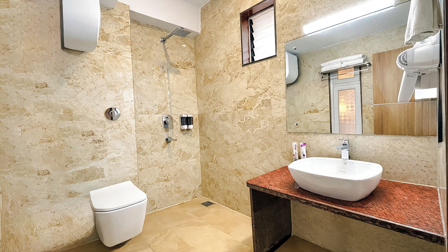 Un baño con pared de mármol, inodoro blanco, ducha, lavamanos con espejo y secador de pelo.