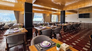 Un ristorante con soffitto e pareti in legno, diversi tavoli apparecchiati con piatti, bicchieri e posate, e un'ampia finestra che dà sul panorama esterno.