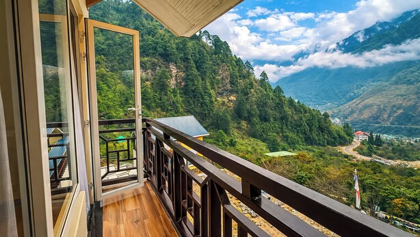 Un balcone con pavimento in legno e ringhiera metallica, affacciato su un paesaggio montuoso sotto un cielo sereno.
