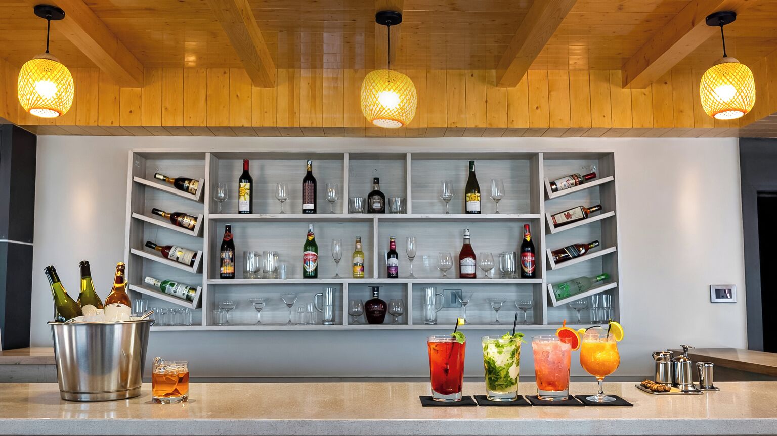A bar area with shelves of bottles and glasses, a bar counter with drinks, and hanging lights.