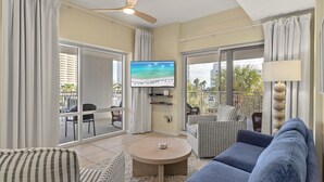 Ein Wohnzimmer mit Fernseher, Sofas, einem Couchtisch und einem Balkon mit Blick auf Palmen und einen Strand.