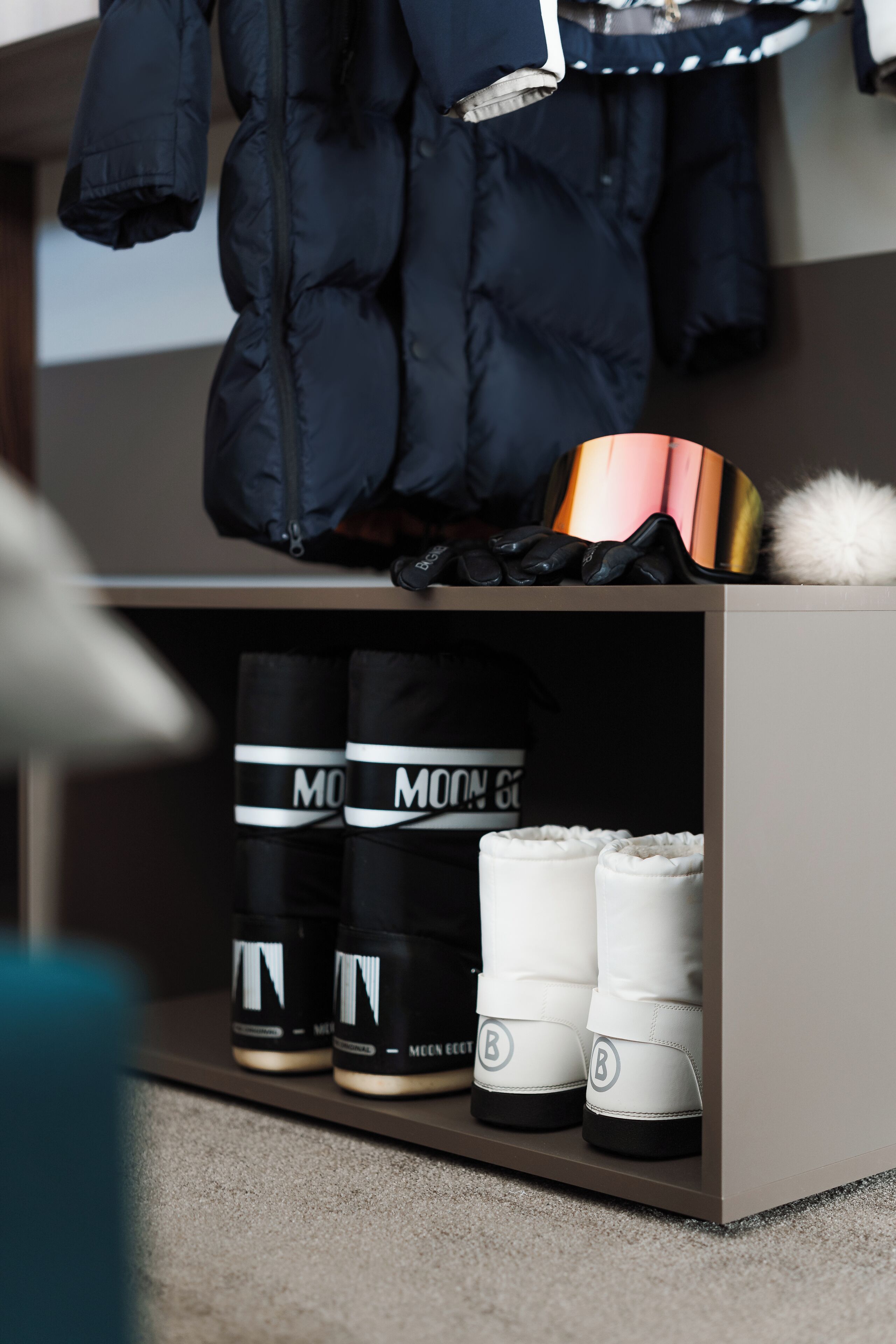 Un módulo de almacenamiento ordenado con zapatillas deportivas blanco y negro, una chaqueta hinchable negra y un objeto blanco y mullido.