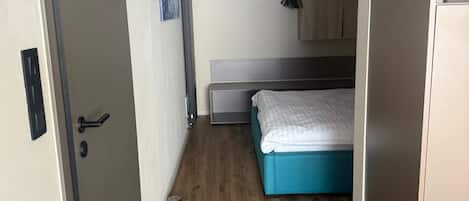 Habitación pequeña con cama, kitchenette y puerta que da a otra zona.
