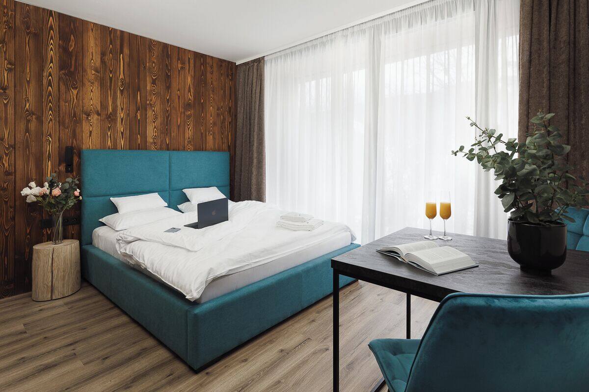 A modern hotel room with a teal bed, wooden panel walls, a wooden floor, a dining table with two chairs, and a potted plant.