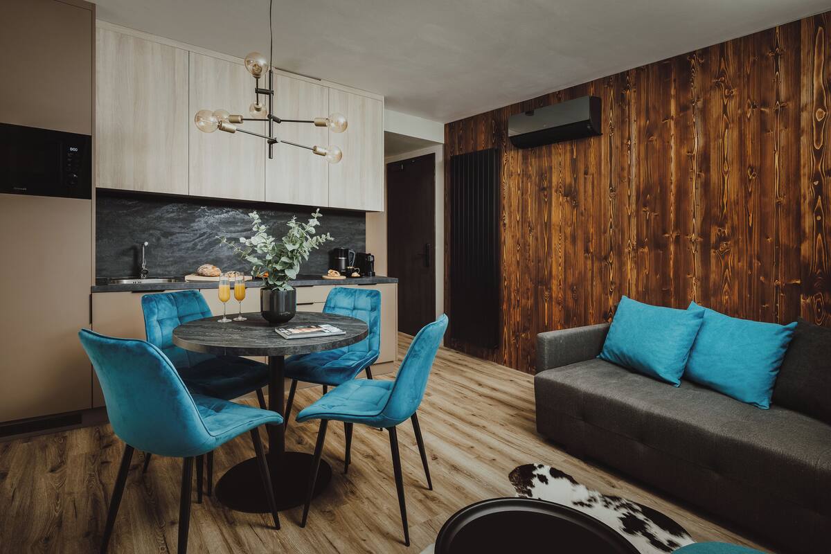 A modern kitchen with a dining area, featuring a round table with blue chairs, a sofa with blue cushions, and a wooden accent wall.