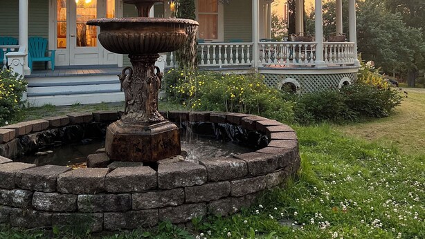 A house with a fountain in the front yard, a porch with chairs, and a sunset in the background.