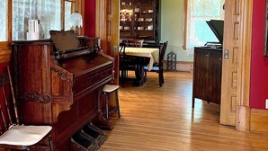 A room with a grand piano, a dining table with chairs, a wooden cabinet, and a large window with curtains.
