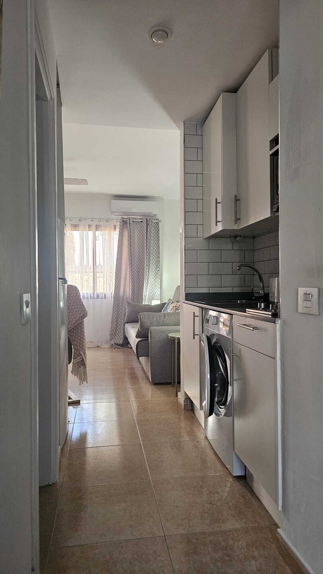 A narrow hallway leading to a kitchen with a washing machine, a dining area with a sofa, and a window with curtains.