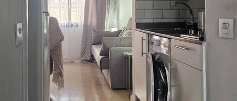 A narrow hallway leading to a kitchen with a washing machine, a dining area with a sofa, and a window with curtains.