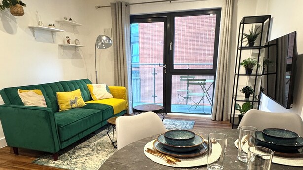 A dining area with a round table set for four, a green sofa with yellow cushions, and a balcony with a view of a brick building. - Modern Canal Side 1BR | Balcony | Near Wembley & Tube (Wembley)
