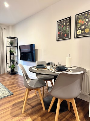 A modern dining area with a round table set for four, surrounded by grey chairs with wooden legs. A black bookshelf with plants is to the left, and a flat-screen TV is mounted on the wall above the table.