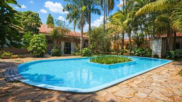A swimming pool surrounded by stone tiles, palm trees, and a building with a red-tiled roof.