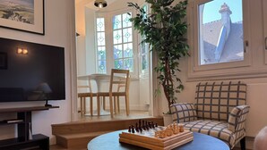 Un salon avec un jeu d’échecs, une télévision, une plante et une fenêtre donnant sur un immeuble.