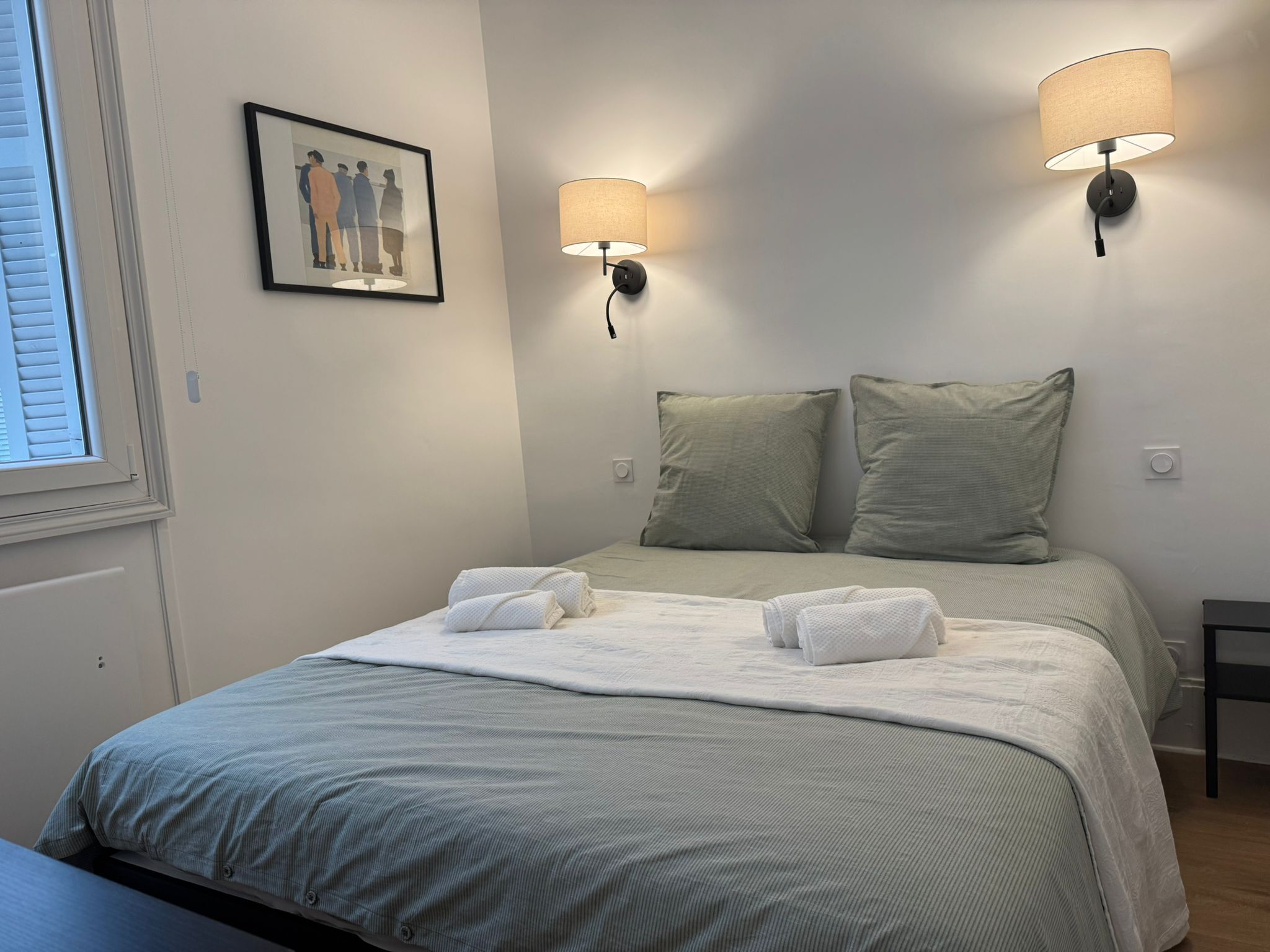 Una cama bien hecha con dos almohadas y dos toallas dobladas, un cuadro en la pared y dos lámparas fijadas a la pared.