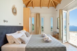A bedroom with a bed, pillows, a bedside table, a mirror, and a view of the outdoors. - Villa Olivia – Panoramic Sea View Retreat (Lefkada)