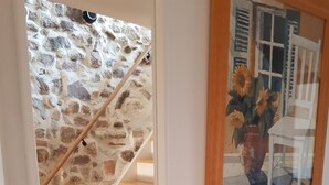 Una escalera con un muro de piedra, un armario blanco con cajones y un cuadro enmarco de una casa con girasoles.