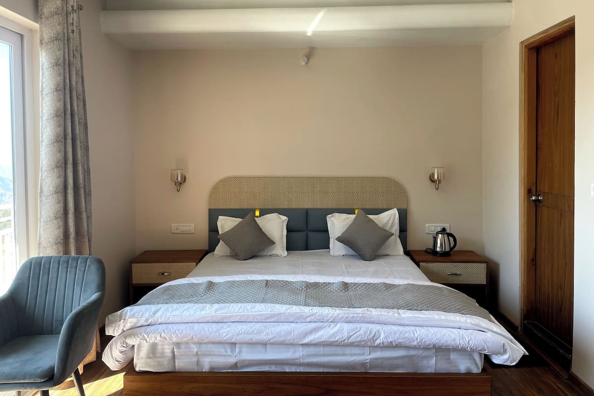 Un dormitorio con una cama, dos mesitas de noche, una silla, una puerta de madera y un ventanal con cortinas.
