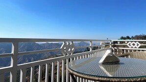 Een balkon met glazen tafel en rieten stoel, met uitzicht op een bergachtig landschap.