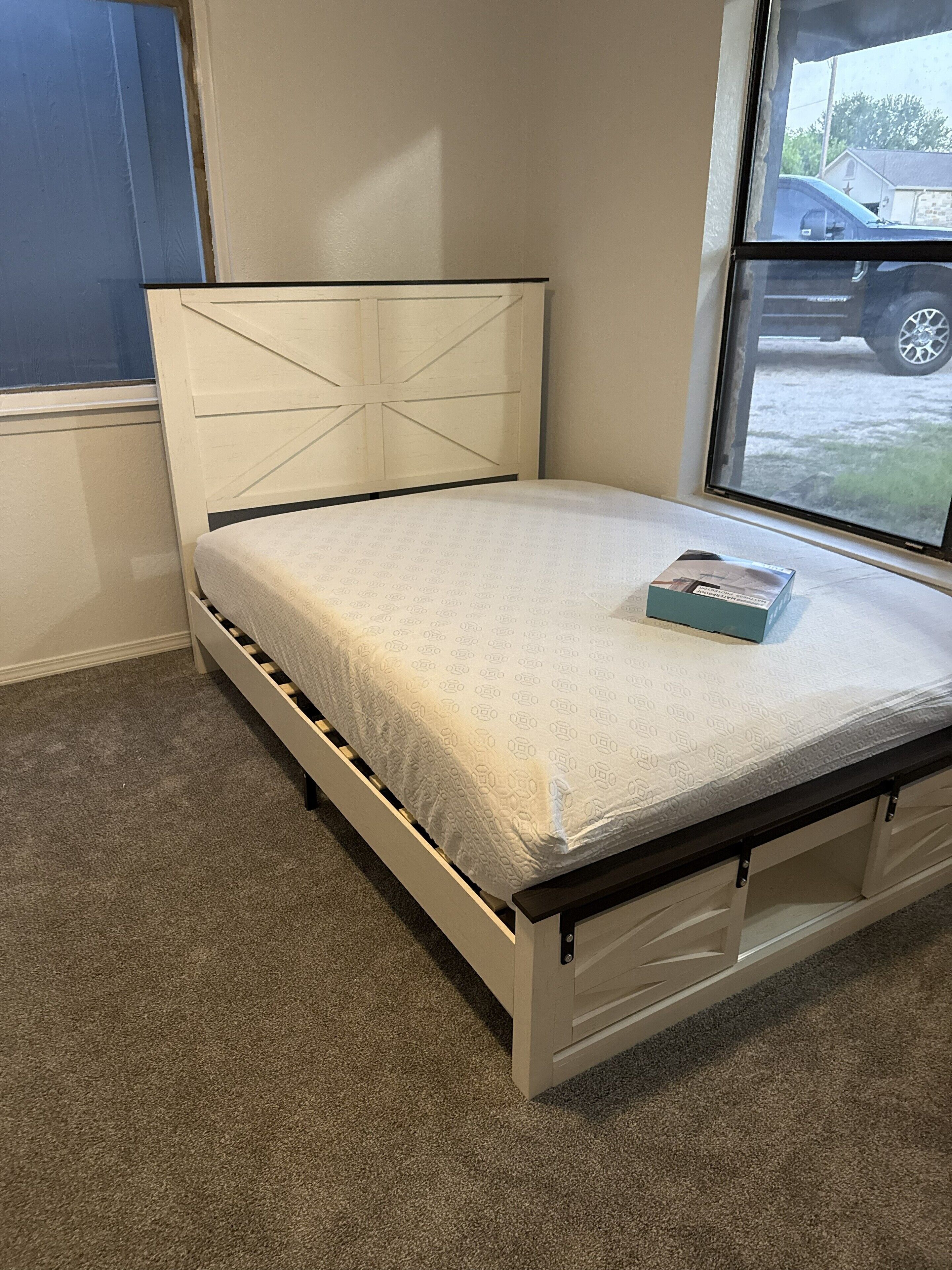 A bed with a white headboard and a mattress with a geometric pattern. There is a window with a view of a parked car outside.