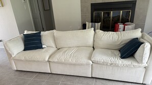 Un canapé d’angle beige agrémenté de deux coussins bleu foncé, un sol carrelé et une cheminée avec une porte en verre en arrière-plan.