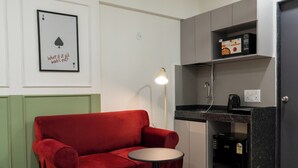 Un espacio habitable compacto con un sofá rojo, una mesita redonda pequeña, una lámpara de pie, una zona de cocina con cafetera y un ventilador de techo.