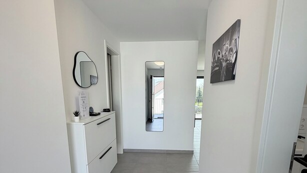 Un couloir avec un mur blanc, un luminaire rond au plafond, une porte donnant sur une autre pièce et un petit tapis sur le sol.
