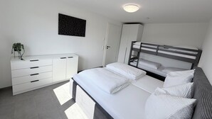 Une chambre avec un lit superposé, une commode blanche, une plante et un luminaire au plafond.