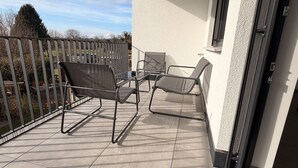 Un balcon avec une chaise en métal, un fauteuil rembourré et une petite table. On peut voir un tapis noir sur lequel est inscrit le mot « BILEANC ».