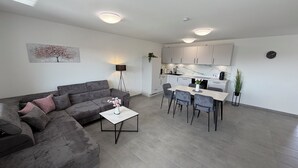 Un salon moderne avec un canapé d’angle gris, une table basse et un coin repas comprenant une table et des chaises. Au fond, on aperçoit une cuisine avec des meubles blancs et un plan de travail en marbre.