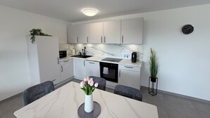 Une cuisine moderne avec des meubles blancs, un plan de travail en marbre, un évier et une machine à café. On y trouve une table à manger ornée d’un vase de fleurs et deux chaises.