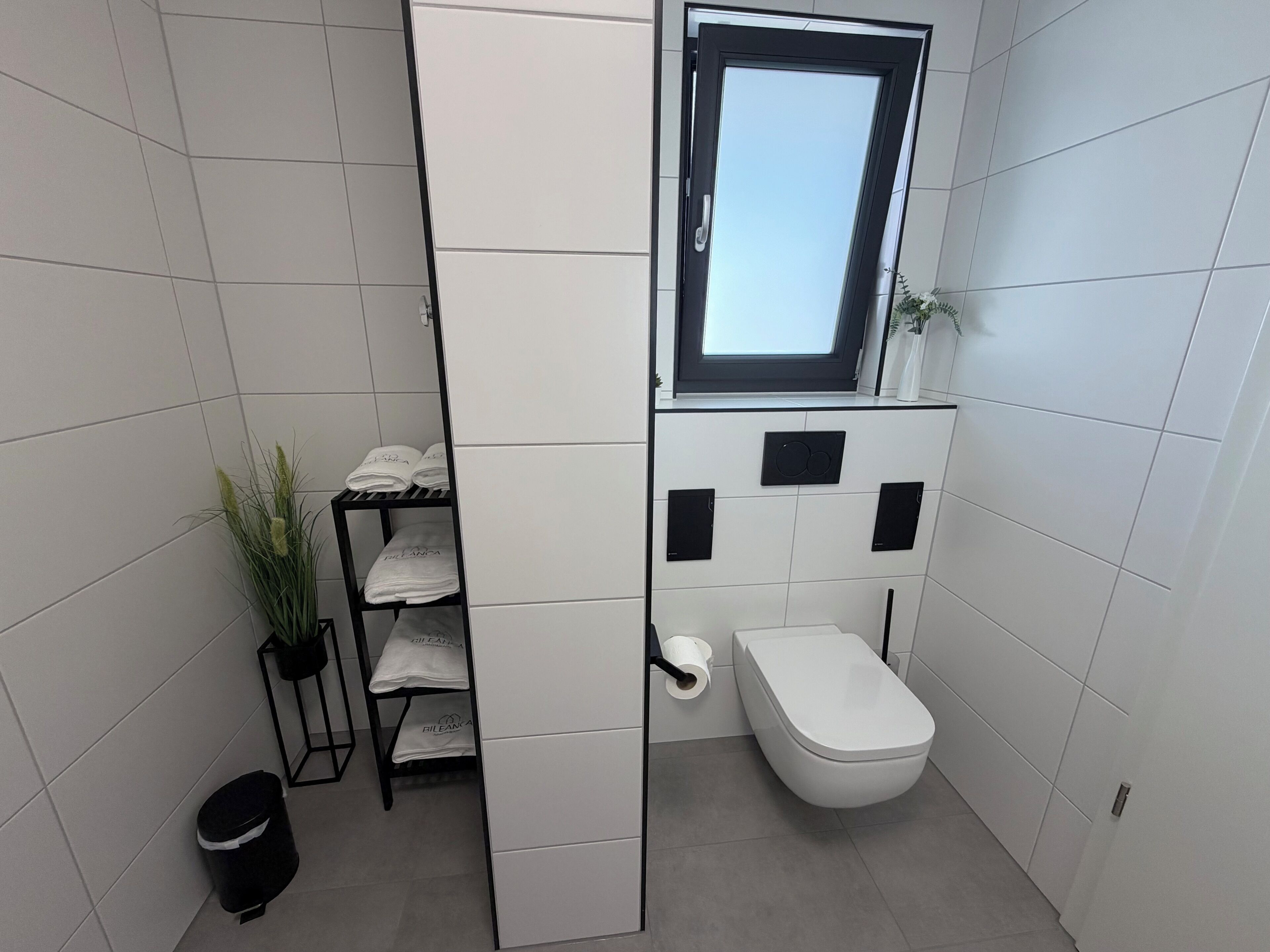 Un baño pequeño y funcional con inodoro, estante con toallas, una planta y ventana.