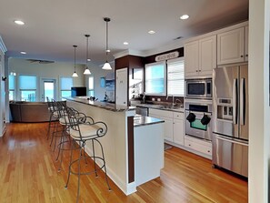 A modern kitchen with a central island, bar stools, stainless steel appliances, and a view of the ocean. - Surf's Up | Oceanfront, Elevator + Prime Location (Carolina Beach)