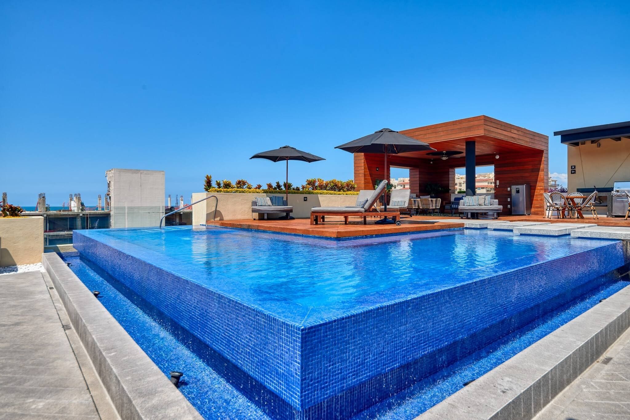 A rectangular swimming pool with tiled edges, surrounded by lounge chairs and umbrellas, and a wooden structure with seating.