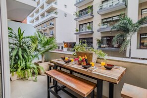 Un balcon avec une table en bois mise pour le petit-déjeuner, entouré de plantes en pot et donnant sur un immeuble moderne.