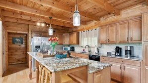 A wooden cabin interior with a kitchen featuring wooden cabinets, a marble countertop, and a central island with stools. - RIVER, HOT TUB, GAME ROOM, PETS - 4 Pool, Playground, Dog Park, Arcade, Tennis. (Ellijay)