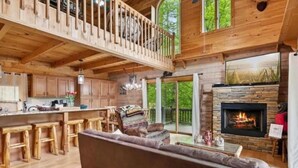A spacious wooden cabin with a high ceiling, a cozy living area with a sofa and a fireplace, and a kitchen with wooden cabinets and a bar area. - RIVER, HOT TUB, GAME ROOM, PETS - 4 Pool, Playground, Dog Park, Arcade, Tennis. (Ellijay)