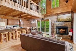 A spacious wooden cabin with a high ceiling, a cozy living area with a sofa and a fireplace, and a kitchen with wooden cabinets and a bar area. - RIVER, HOT TUB, GAME ROOM, PETS - 4 Pool, Playground, Dog Park, Arcade, Tennis. (Ellijay)