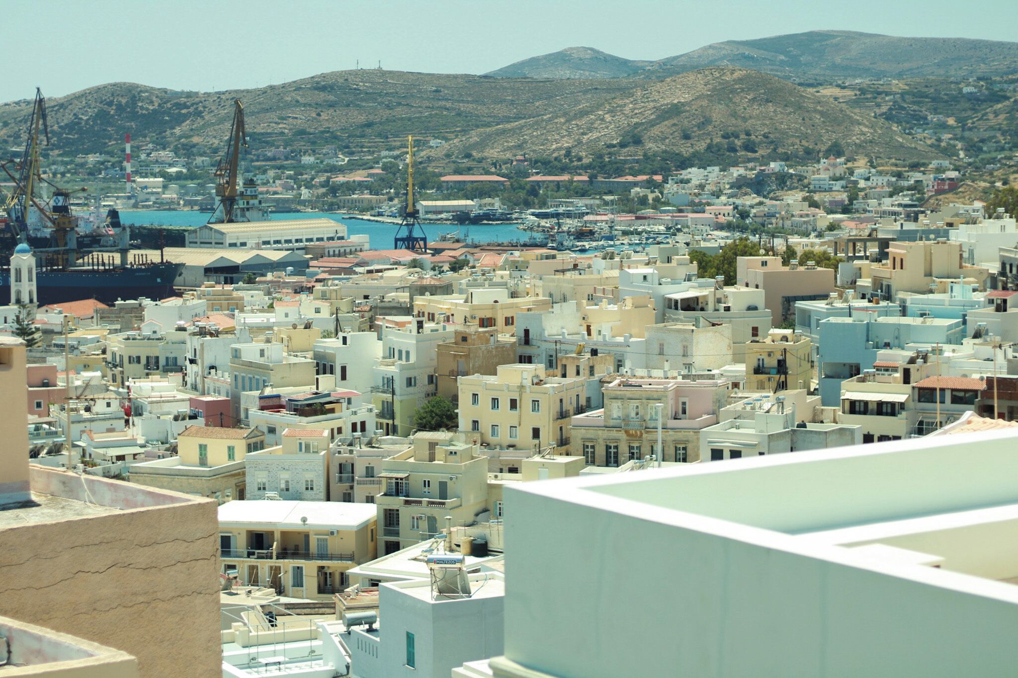Un paysage urbain côtier avec un port, comprenant un grand navire et des grues, ainsi que des bâtiments densément regroupés présentant une architecture variée.