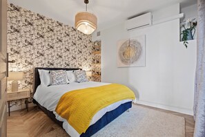 A bedroom with a bed, a bedside table, a lamp, a wall with a floral wallpaper, and a ceiling light. - Baie des Anges (Nice)