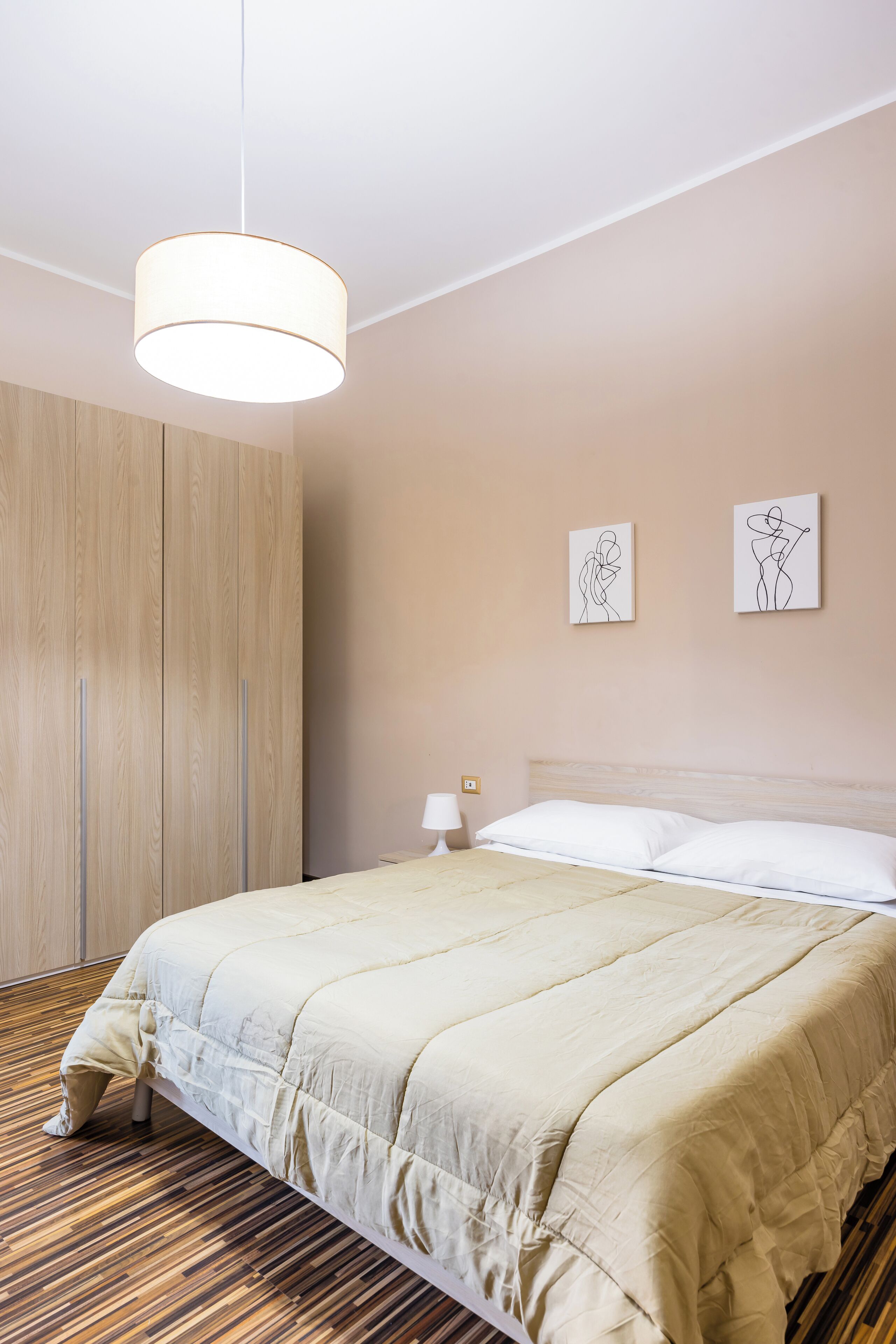 A neatly made bed with a beige comforter, a wooden wardrobe, and two wall art pieces depicting abstract figures.