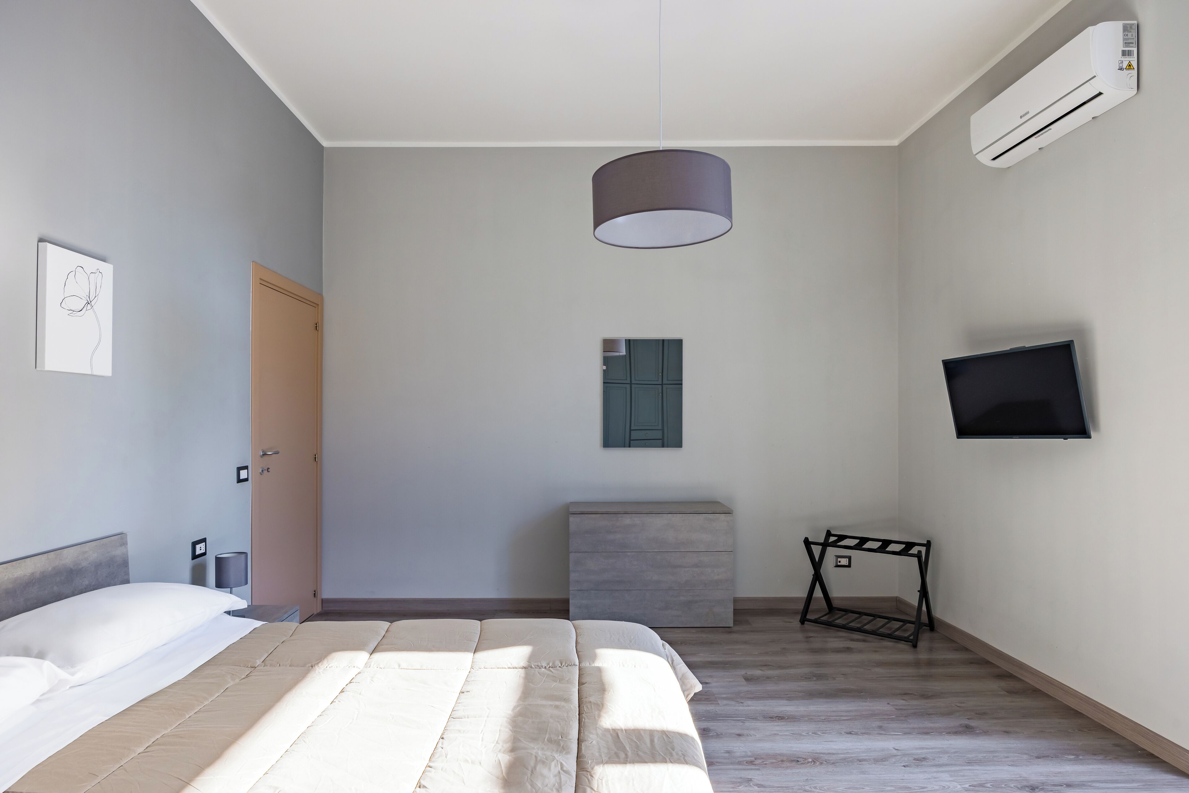 A modern bedroom with a bed, bedside table, lamp, air conditioning unit, TV, and a small cabinet.