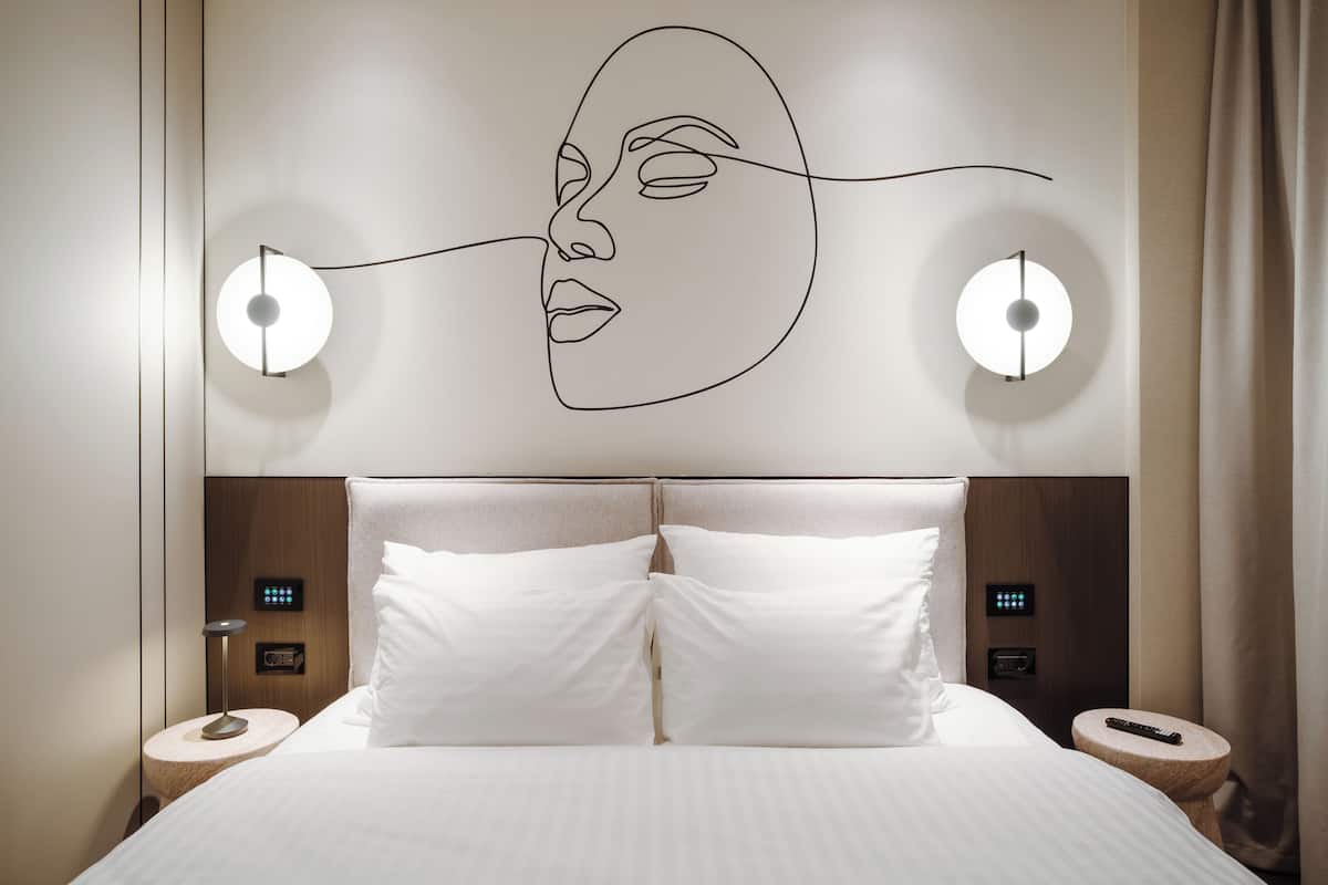 A hotel room with a large bed, two bedside tables, a wall-mounted light fixture, and a control panel on the headboard.