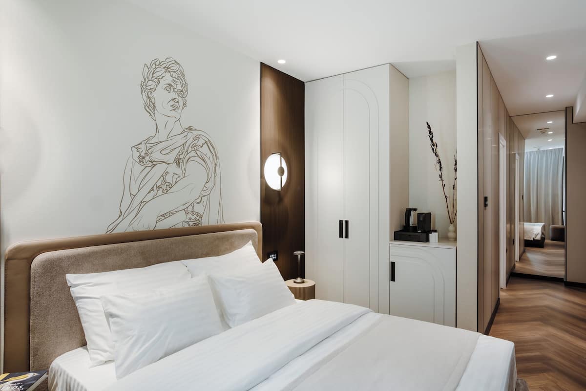 A modern hotel room with a large bed, a wall-mounted artwork, a wooden accent wall, a bedside table with a lamp, and a wardrobe.