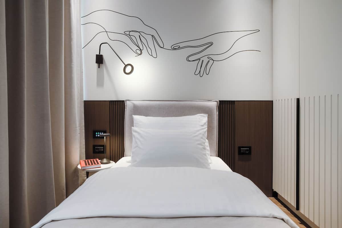A modern hotel room with a large bed, a bedside table with a lamp, and a wall art of hands reaching towards each other.