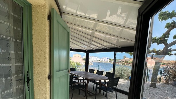 Eine überdachte Terrasse mit Tisch und Stühlen, eine grüne Tür und Blick auf ein Gewässer.