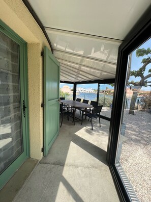 Un patio techado con una mesa y sillas, una puerta verde, y vista hacia un cuerpo de agua.