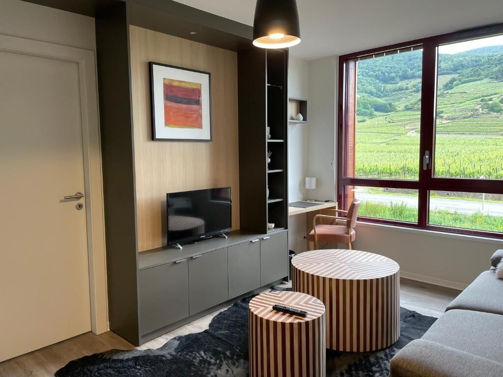 Een moderne woonkamer met een televisie, twee gestreepte voetenbankjes, een bank en een groot raam met uitzicht op groene velden.
